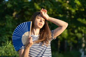 Young woman suffering heatstroke outdoors using fan to get fresh air. Unhappy girl feel bad of hot...