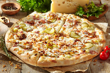 Tasty pizza with ham, gherkins, fried onions and pickle cucumber. Garnished on parchment with pizza ingredients on wooden table. Rustic style, sunlight.
