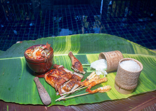 .Sticky rice, papaya salad, grilled chicken and grilled shrimp served on a spotted banana leaf poolside..red yellow green speckled banana leaves in the background. food and restaurant concept...