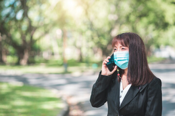 business woman wearing face mask using smartphone tablet in calling for working with her customer in park with sunrise and blur nature background. business new normal with corona virus or covid-19.