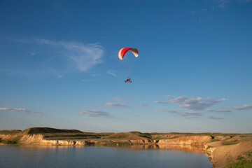 A red paraglider flies in the blue sky over the lake and the picturesque steep coast of the concept of extreme sports.