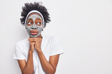 Surprised pensive woman with curly hair keeps hands under chin looks away applies clay mask for reducing fine lines wears casual t shirt isolated over white background blank copy space for your text