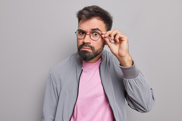 Serious bearded man keeps hand on rim of spectacles has attentive gaze directly at camera has...