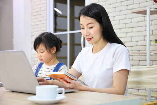 Asian mother working online from home using laptop while watching her daughter working on assignment from school for homeschooling and education concept
