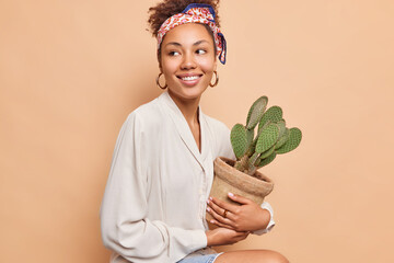 Pretty dark skinned curly haired woman sits against beige background looks happily away holds pot ot prickly succulent cactus for her home garden wears fashionable clothes isolated. Houseplants