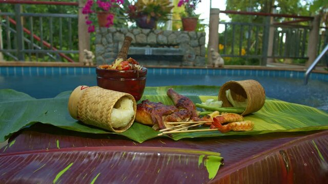 Sticky rice, papaya salad, grilled chicken and grilled shrimp served on a spotted banana leaf poolside..red yellow green speckled banana leaves in the background. food and restaurant concept.