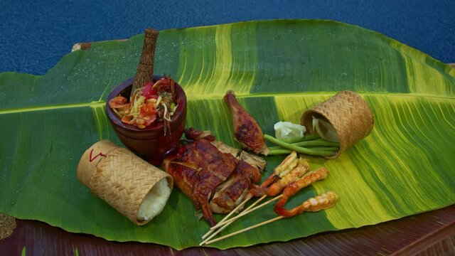 Sticky rice, papaya salad, grilled chicken and grilled shrimp served on a spotted banana leaf poolside..red yellow green speckled banana leaves in the background. food and restaurant concept.