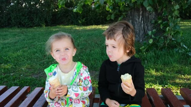 Cute little children eat delicious ice cream sitting on bench in green park. Brother and sister enjoy cold dessert on spring day