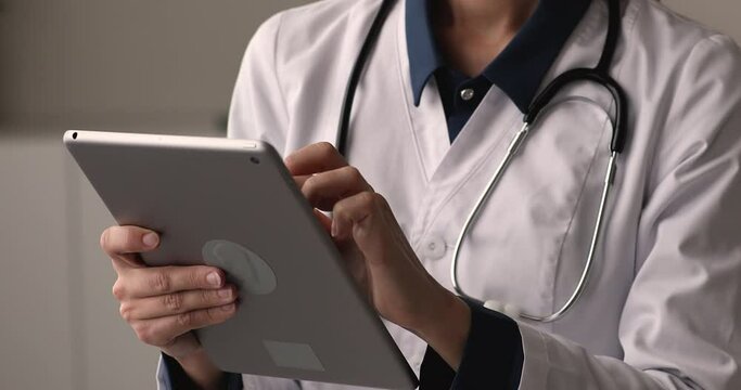 Close up young female general practitioner doctor using digital tablet, working distantly typing web surfing information, checking daily agenda, managing meeting, modern technology apps for medicine.