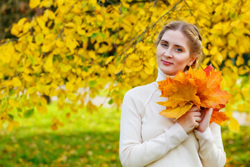 Woman in an autumn park. An emotional portrait of a cute and positive beautiful blonde girl with a bouquet of maple leaves on a background of blurred yellow foliage