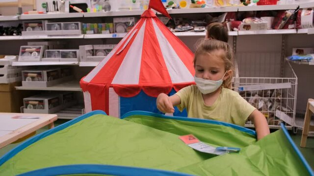 Blonde little girl in medical mask plays with green tent while boy walks past shelves in toy store. Shopping during quarantine