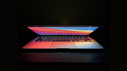 Laptop with half  lid open on a table lit with colorful desktop screen wallpaper in a dark room