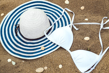 white women's swimsuit lies on the sand with seashells along with a sun hat.