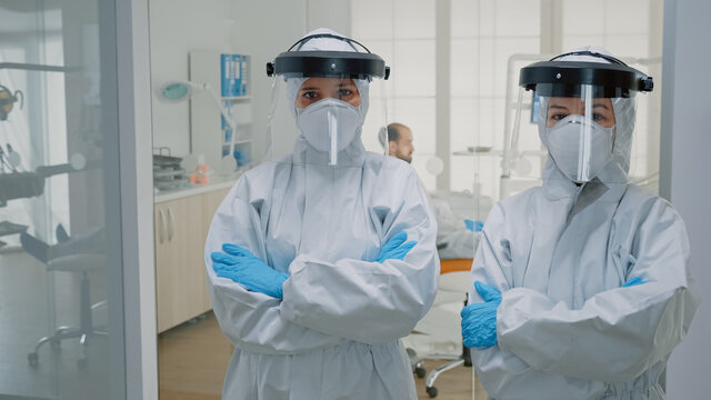 Professional team of dentists in ppe suits standing in dental cabinet looking at camera. Stomatologists wearing virus protection uniform preparing for oral patient examination