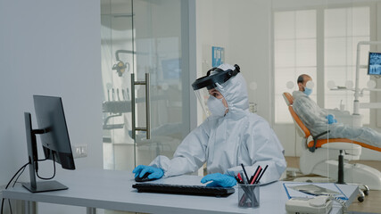 Stomatology team of dentists at desk using computer for teethcare in dental cabinet, wearing virus protection suits. Nurse with modern device sitting while orthodontist preparing for patient