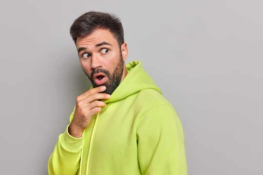 Startled bearded young man holds chin opens mouth from amazement stares speechless in awe cannot believe his eyes wears comfortable green hoodie poses against grey background copy space on right