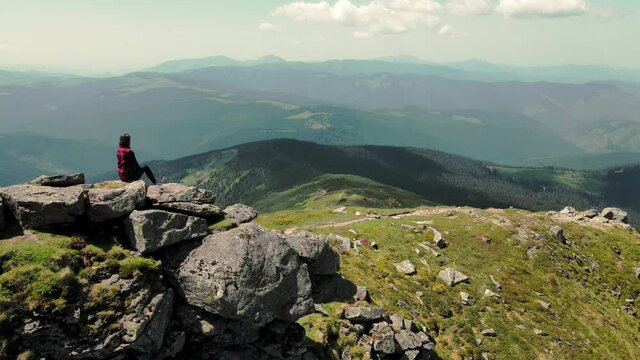A woman sitting on top of a mountain looks into the distance at the beauty of the Carpathian mountains. Aerial view. Silhouette of a woman sitting on a rocky mountain. View of a young girl from the