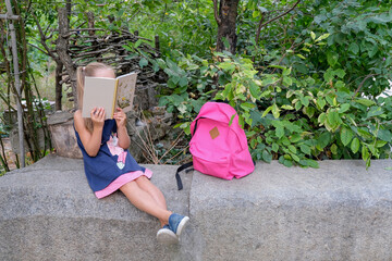Back to school. A girl with school backpacks sits at recess in a school park and reads a book. Prepare for the lesson. The child walks near the school. New year students.
