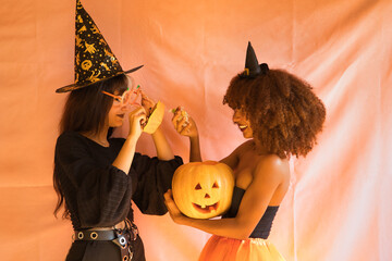 Young Hispanic and Latina women dressed as witches pulling candy out of a pumpkin on a pink...