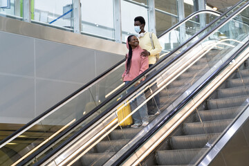Happy african american couple travel in covid-19 lockdown. Black man wearing medical mask hug woman holding suitcase on escalator arriving from flight. Tourism and transport in coronavirus quarantine