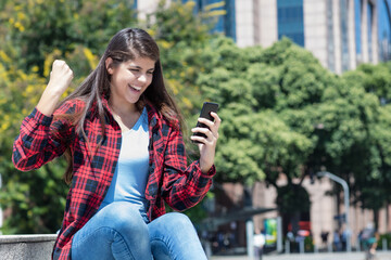 Cheering spanish young adult woman receiving message with good news on phone