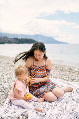 Mom feeds little girl a banana while sitting on the beach