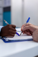 Close up of hands holding pen on document files for signature at healthcare office desk. Caucasian senior man signing appointment papers for checkup with african american doctor