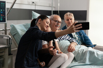 Caring family taking selfie with sick elderly grandfather using modern smartphone during clinical recovery in hospital ward. Relatives visiting senior man support him after medical surgery