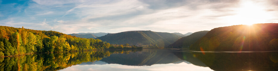 Obraz premium beautiful autumn landscape by the lake at sunset. panorama with trees in colorful foliage and forested apuseni mountains reflecting in the water surface. clouds on the sky in evening light