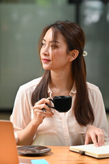 Portrait with Asian businesswoman  sitting on her office desk and holding a cup of coffee while looking out side the window.