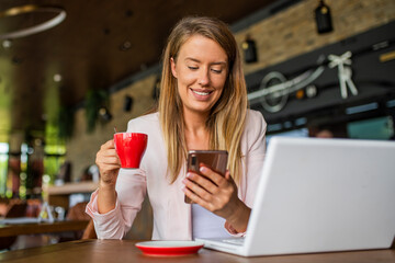 Attractive young woman holding coffee cup and looking at the laptop in restaurant. Beautiful girl with smart phone in cafe. Young woman using smartphone and laptop