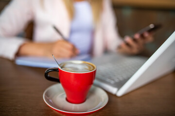 Business woman indoor with coffee and laptop taking notes.  Young woman sitting in coffee shop at wooden table, drinking coffee and using smartphone.On table is laptop. Girl browsing internet