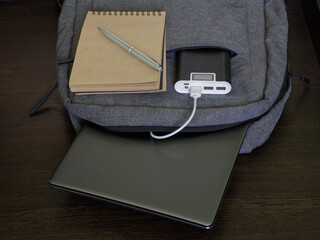 Modern laptop backpack sits on a wooden table along with a silver laptop, power bank, notepad and pen. The concept of recharging gadgets while studying, traveling and trip