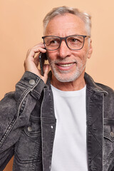 Bearded smiling grey haired man dressed in fashionable black jacket has phone talk looks into...