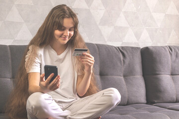 Positive woman with credit card and mobile phone in hand sitting on sofa at home. Making online order or purchasing goods on internet concept background