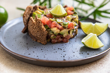 Raw tuna and avocado tartar served with rye bread crackers and lime on black ceramic plate. Gourmet fish tartar.