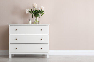 White chest of drawers with bouquet and candle near beige wall. Space for text - obrazy, fototapety, plakaty