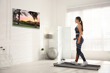 Fototapeta na wymiar Sporty woman training on walking treadmill and watching TV at home