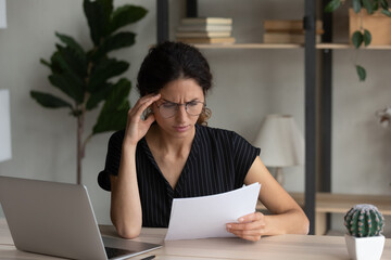 Close up dissatisfied businesswoman in glasses reading bad news in letter, checking financial documents, sitting at work table with laptop, confused stressed woman worried about money problem or debt