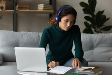 Close up focused woman in headphones studying online, taking notes, reading book, using laptop, listening to lecture or watching webinar at home, student learning language, involved in internet class