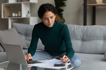 Focused woman using laptop and calculator, checking domestic bills, sitting on couch at home,...