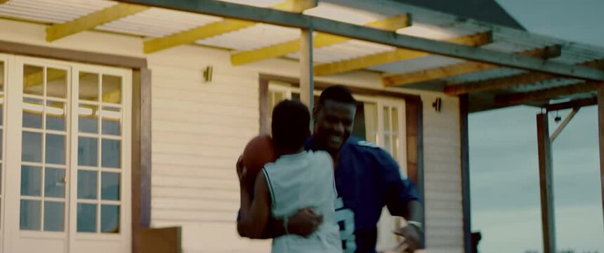 HANDHELD African American father and son playing football together on a lawn in front of their house in the evening. Shot with 2x anamorphic lens