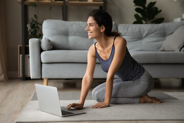Smiling dreamy woman in sportswear using laptop, waiting for online training, sitting on floor in...