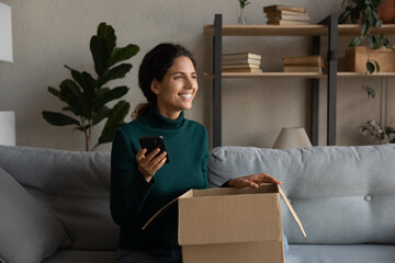 Smiling woman excited by online store order, holding smartphone, opening cardboard box, sitting on...