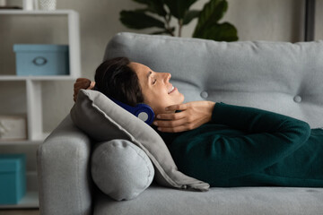 Close up profile of smiling woman in headphones enjoying music, relaxing, lying on soft pillow, positive attractive young female with closed eyes listening to favorite song, resting on couch at home