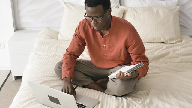African-American guy with glasses in pajamas works holding pen on laptop sitting on comfortable bed in light room closeup