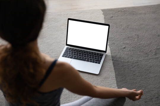 Top view over shoulder woman practicing yoga online at home, laptop with blank empty white screen mockup, young female involved in online lesson with trainer or watching webinar, meditating