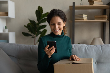 Close up smiling young woman using phone, checking online order, cardboard unpacking box, sitting...