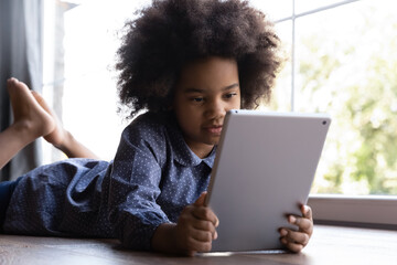 Cute smart little teenage African American girl child lying on floor at home talk speak on video webcam call on pad. Small teen ethnic biracial kid have fun using modern tablet gadget studying online.