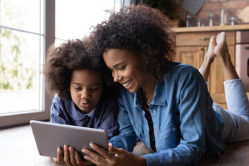 Happy loving young African American mom and teen daughter relax on wooden kitchen floor have fun using tablet together. Smiling ethnic mom and biracial girl child speak on webcam video call on pad.
